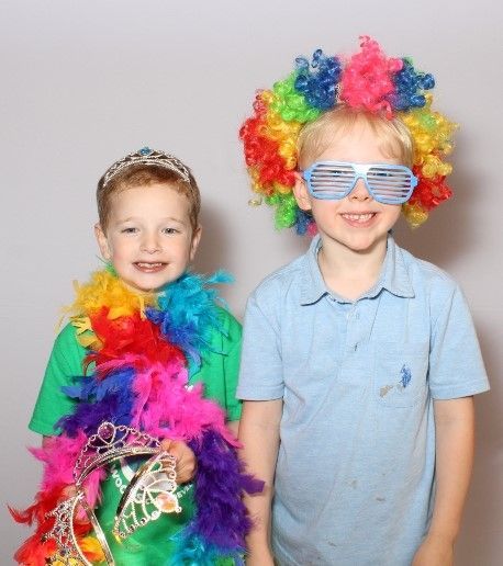 Two young boys in costume, one wearing a rainbow boa and tiara, the other a rainbow wig and shutter shades, smiling.