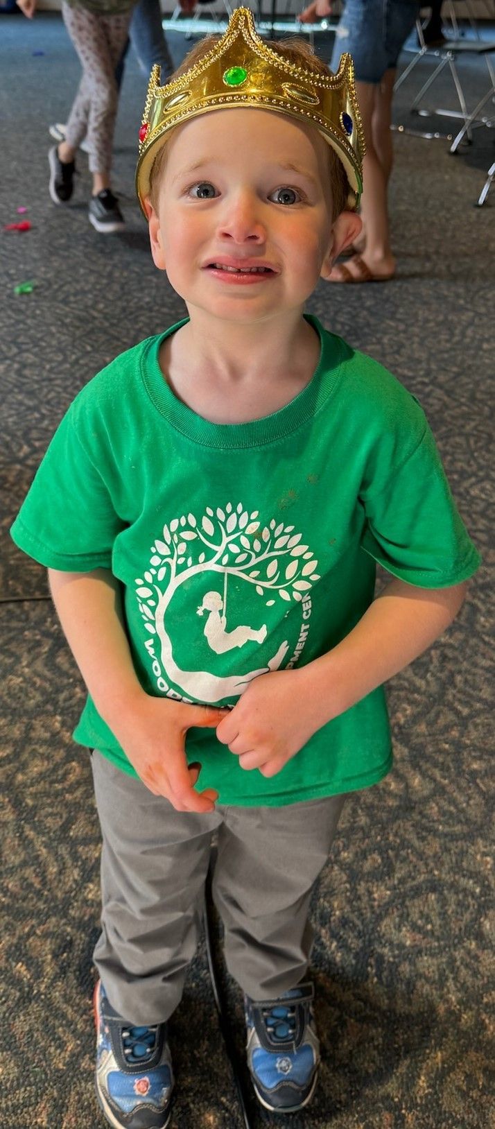 A young child wearing a gold crown and green shirt with a white logo, gray pants, and blue shoes stands inside. The child appears to be slightly sad.
