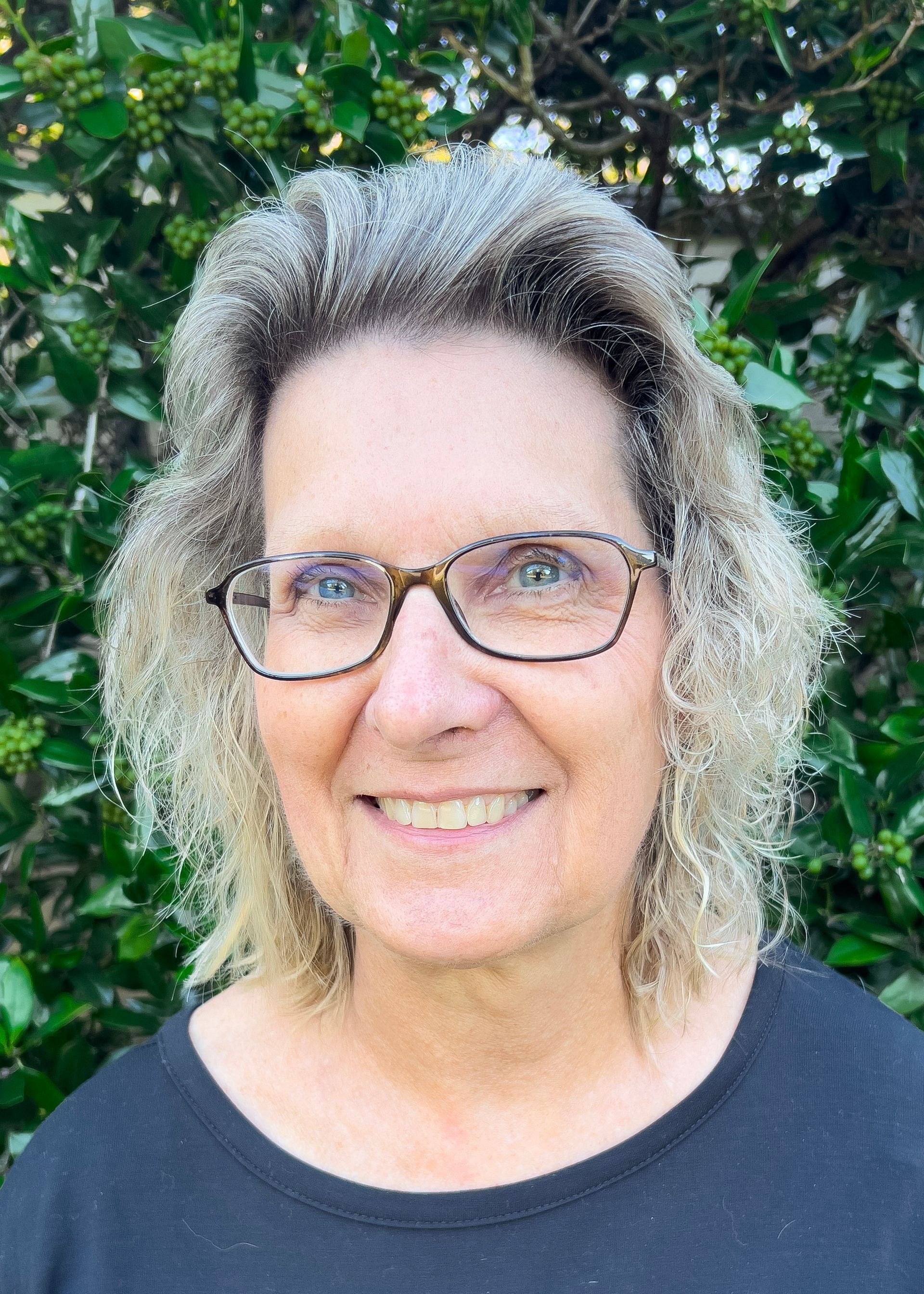Woman with glasses smiles outdoors; gray and blonde hair; black shirt.