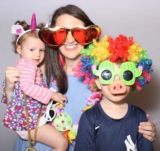 Woman and two children wearing silly photo booth props: heart-shaped sunglasses, a rainbow wig with cactus glasses and a pig nose, and a unicorn headband.