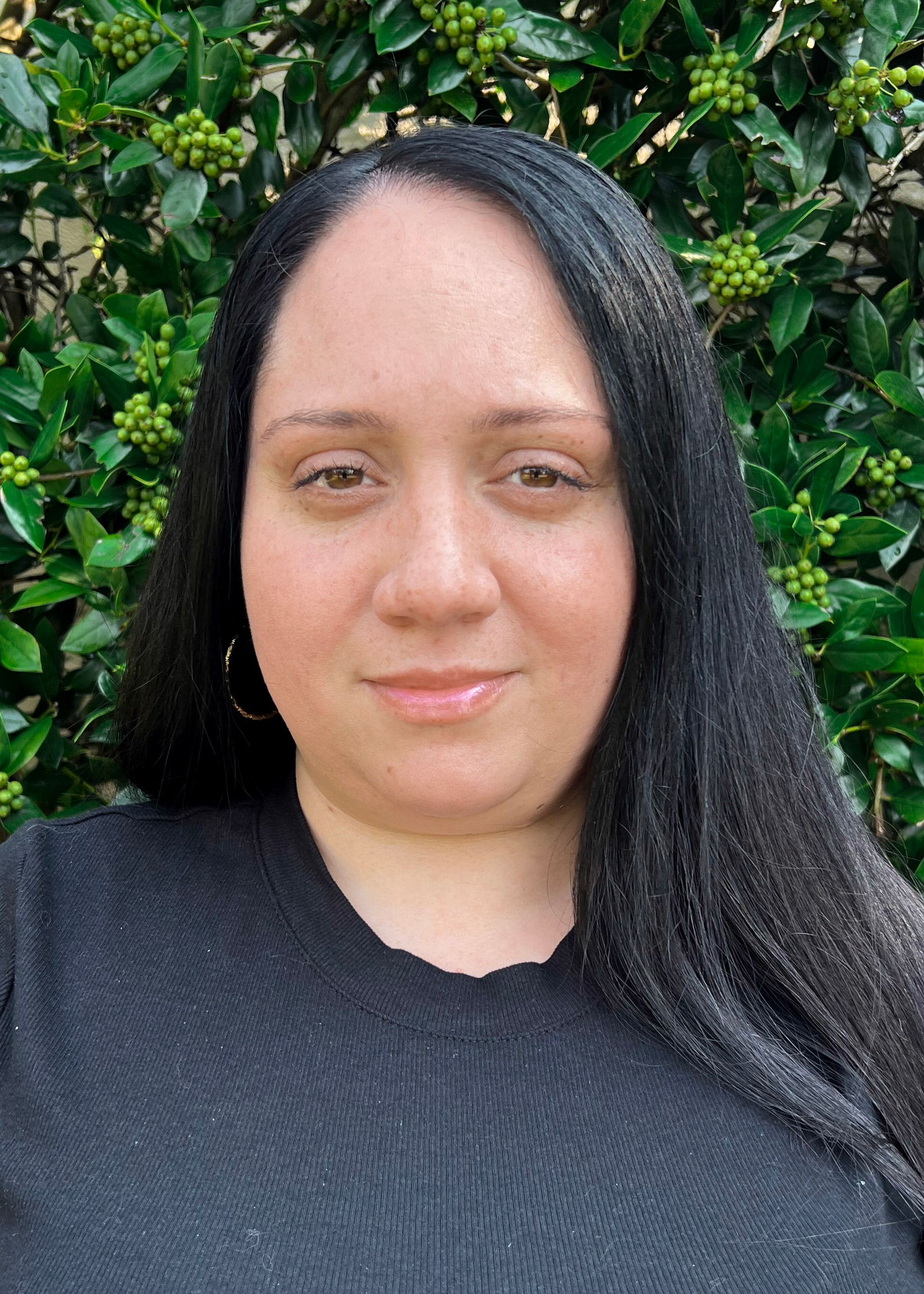 Woman with long black hair wearing a blue denim jacket, standing in front of a tree with brown bark and green leaves.