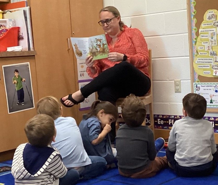 A woman reads a book to a group of young children sitting on a blue mat in a classroom.