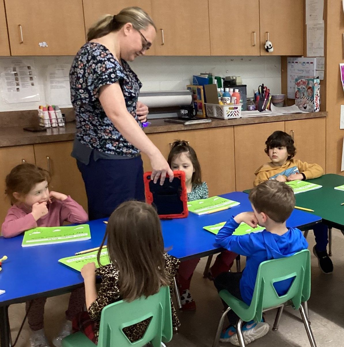 A teacher shows a tablet to five young students seated at a blue table in a classroom setting.