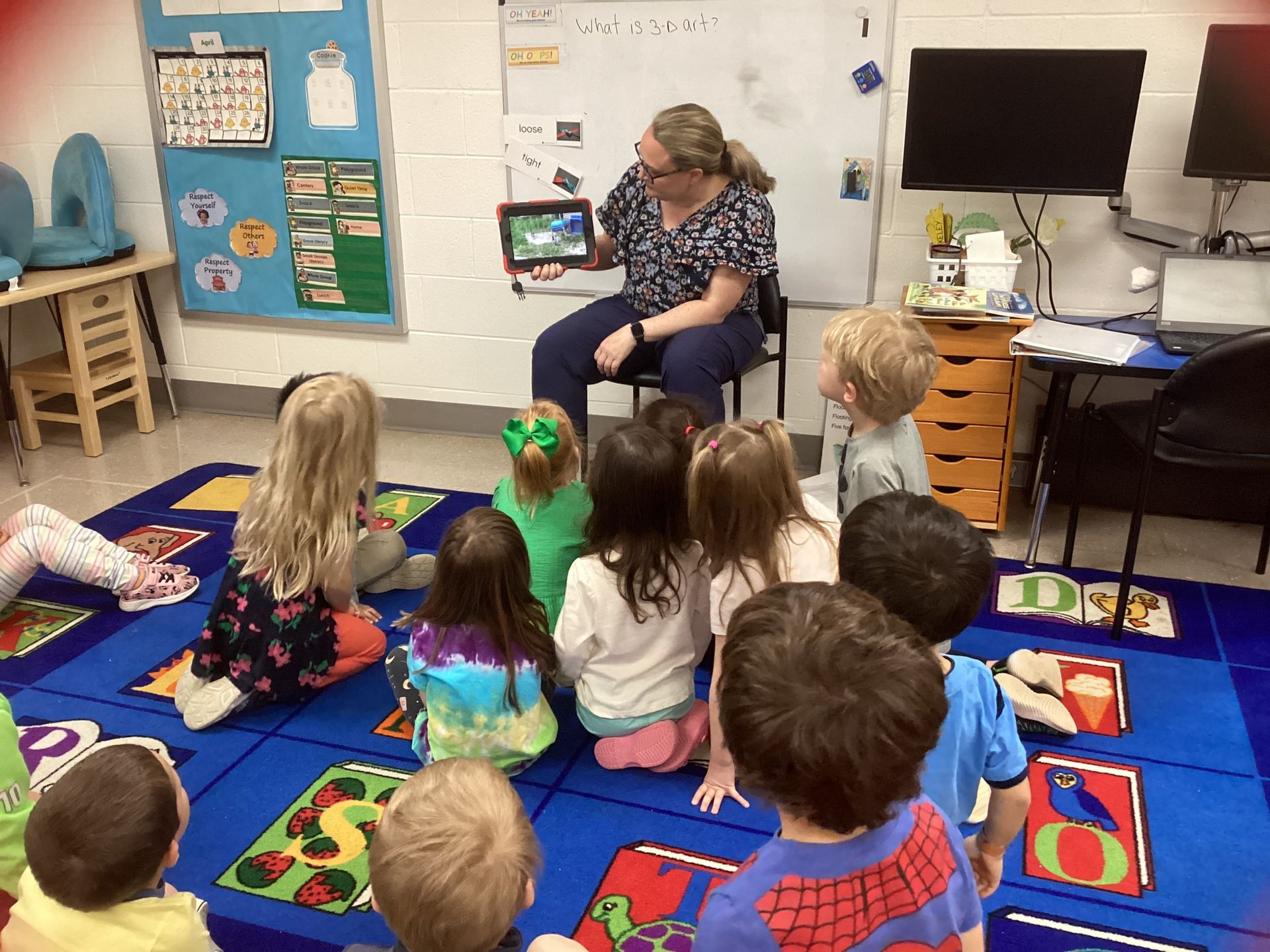 Teacher shows tablet to a group of young children seated on a blue rug in a classroom.