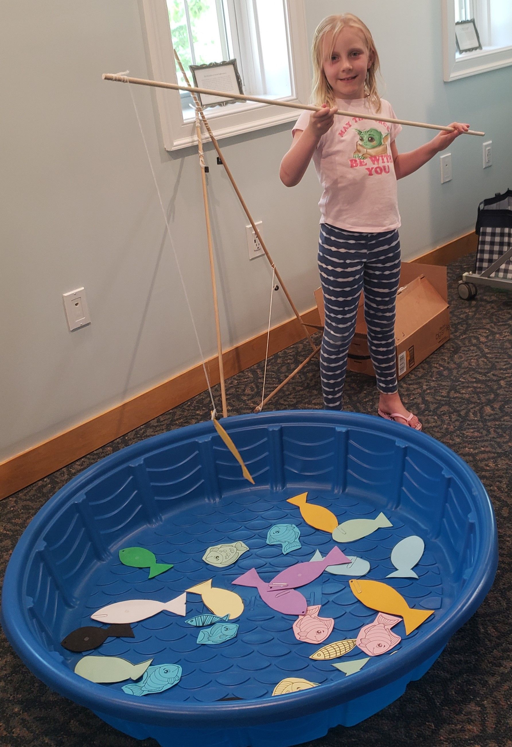 A young girl smiles while holding a fishing rod over a blue plastic pool filled with colorful paper fish.