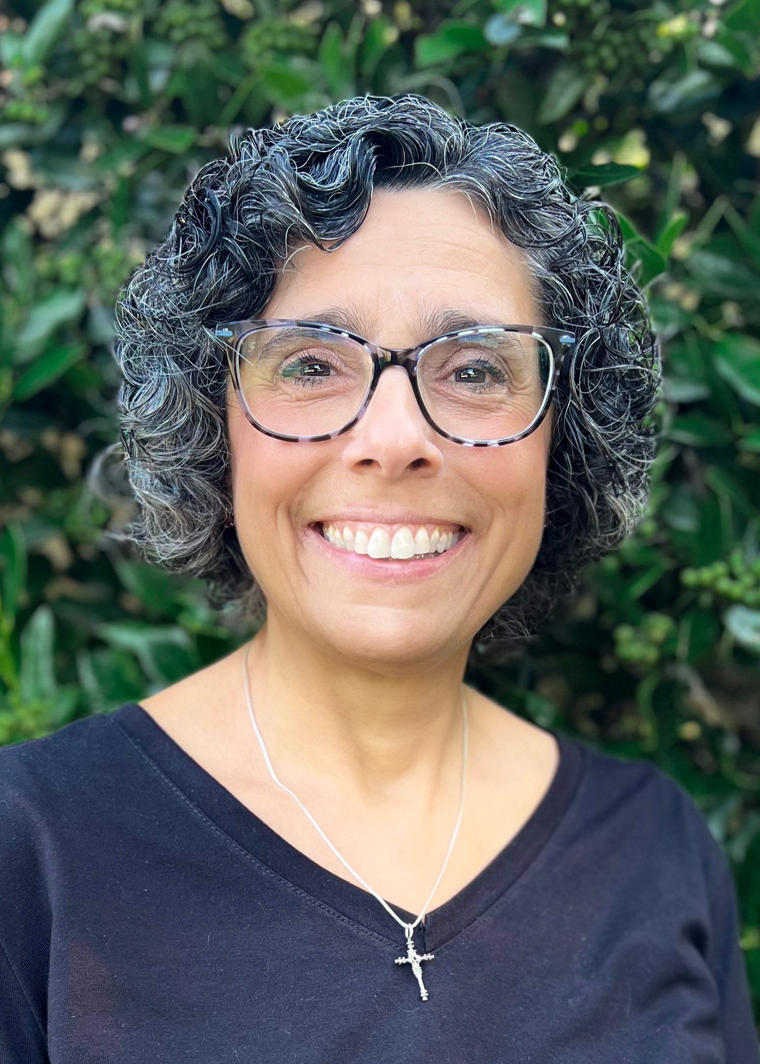Woman with glasses smiles, dark hair, black shirt, necklace with cross; set against green foliage.