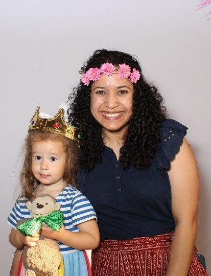 Woman with flower crown smiles beside child wearing a crown and holding a monkey. They stand in front of a white background.