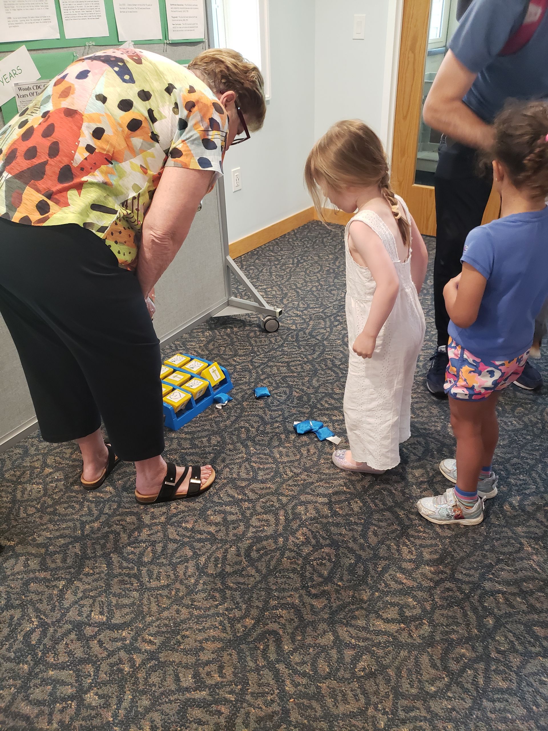 A woman and two young children looking at toys on a carpeted floor. The woman bends over; the children watch with interest.