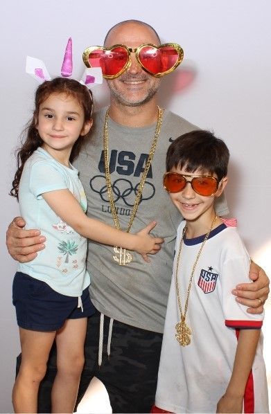 A man with novelty glasses, and two children pose; the man is wearing a gold chain, and the girl has unicorn horns.
