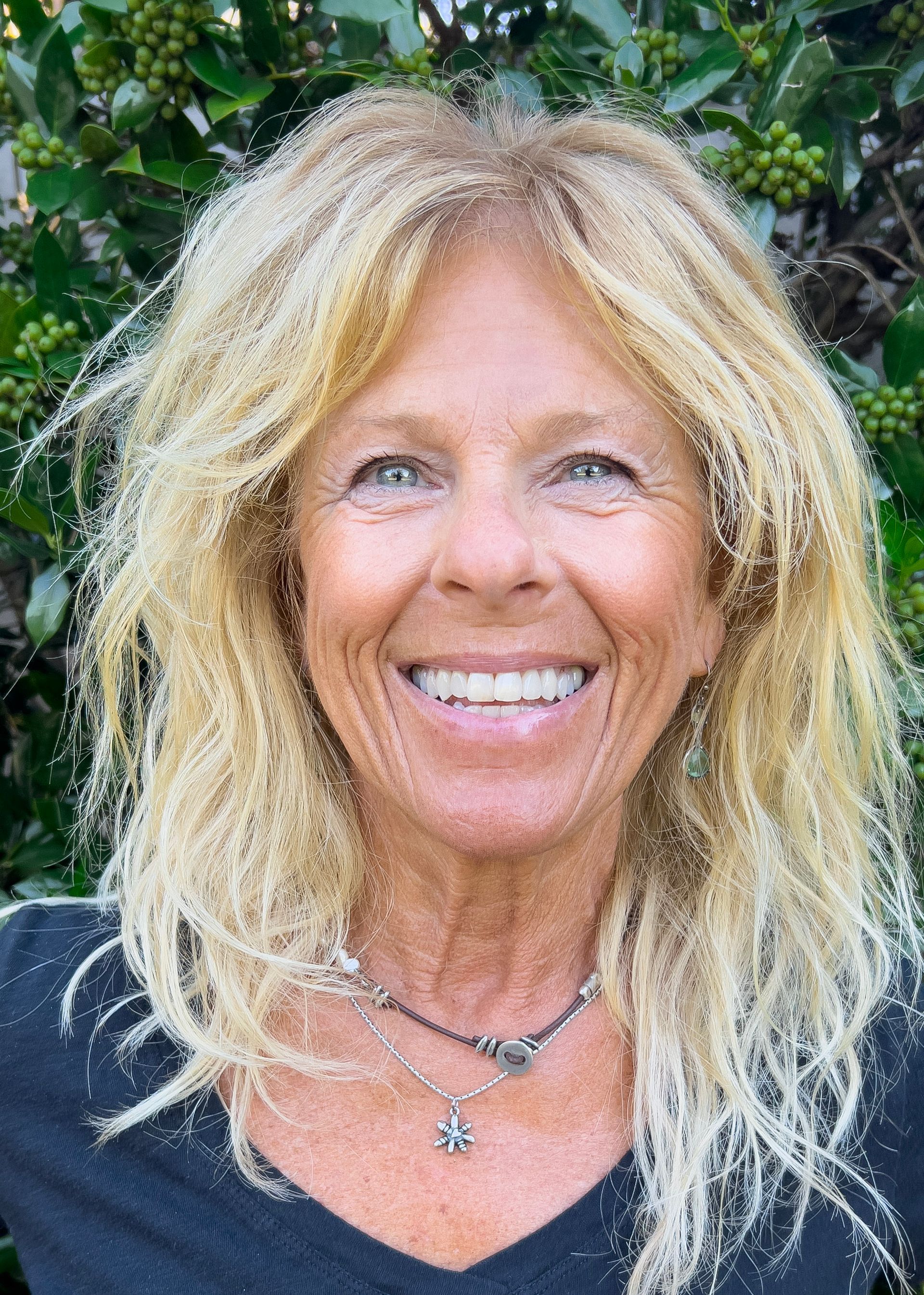 Blonde woman smiling, wearing a dark shirt and necklace, in front of green foliage.
