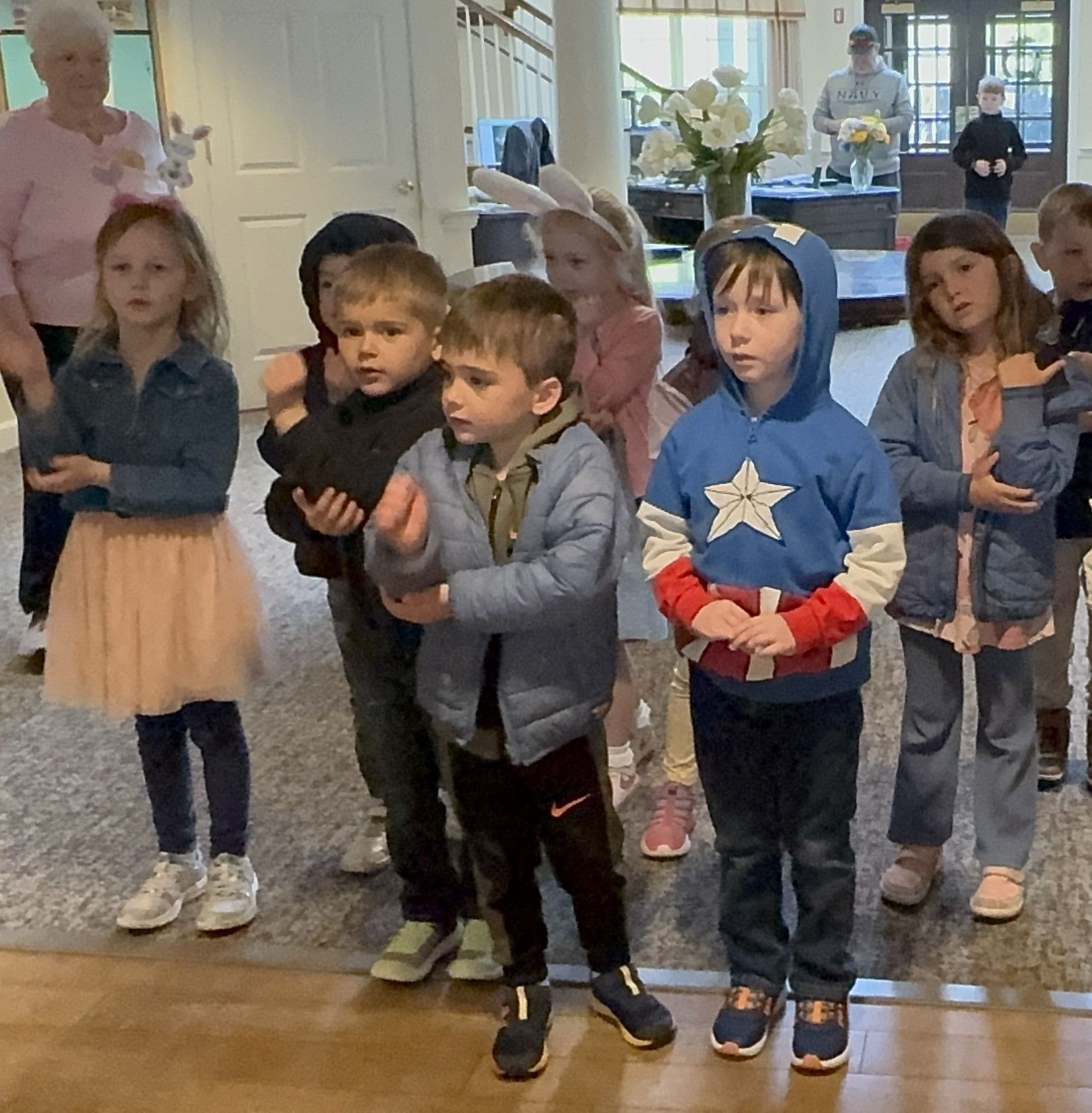 A group of children in casual clothes and bunny ears stand with their hands up while looking at the camera. A woman and a man are in the background.