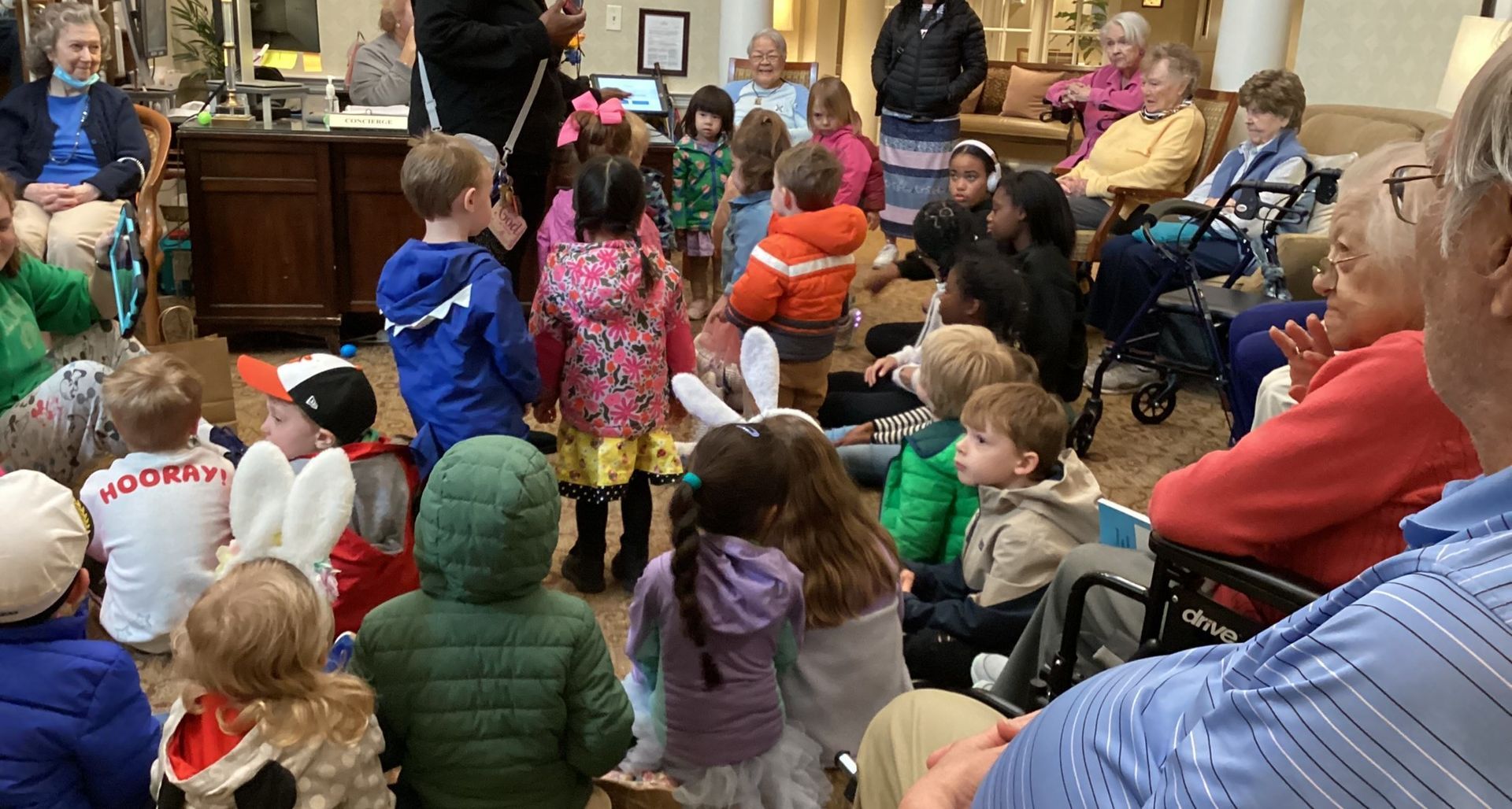 Children dressed in spring attire perform for an audience of senior citizens in a common area; some wear bunny ears.