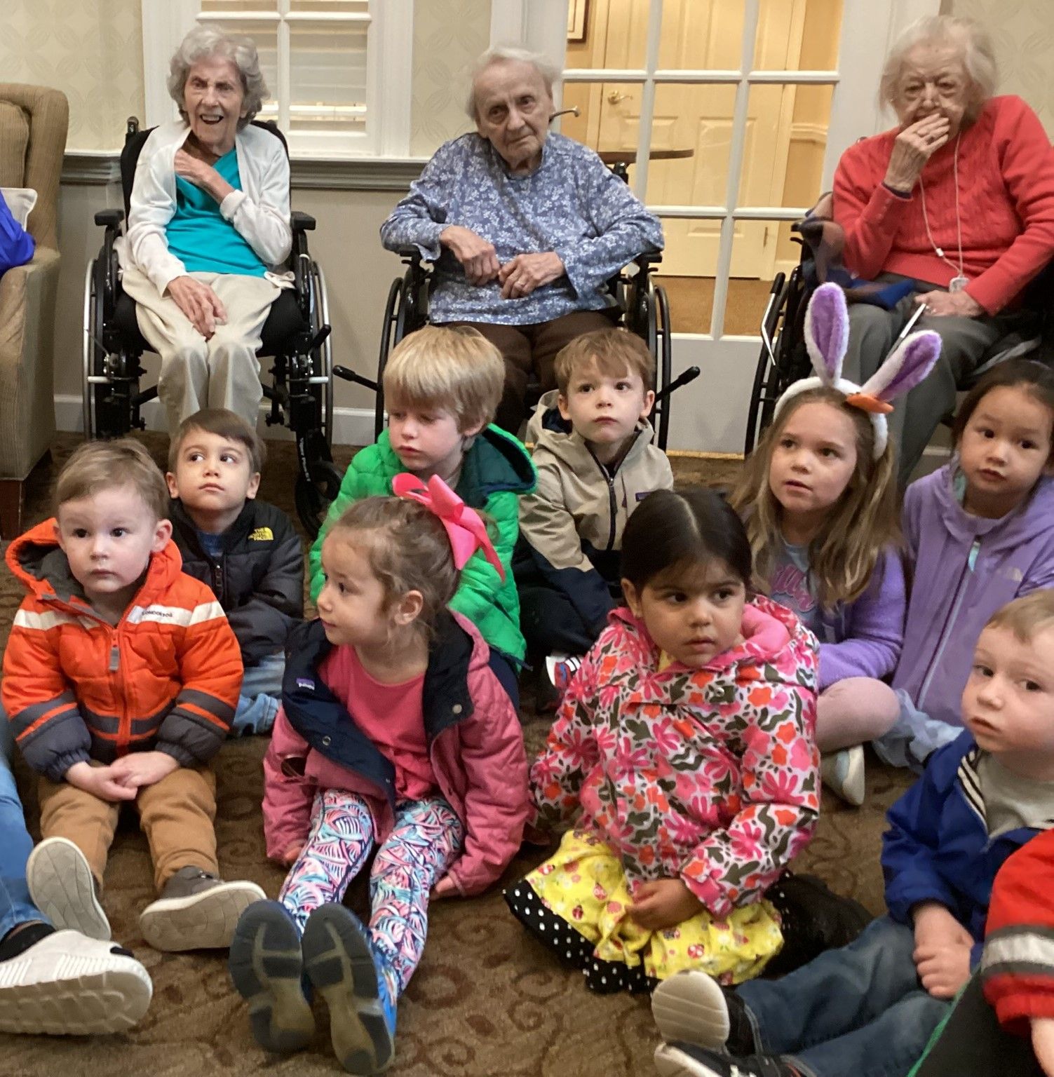 A group of children sit on the floor facing three elderly women in wheelchairs. The group is indoors.