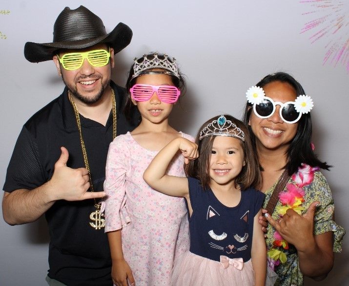 Family in photo booth wearing props: cowboy hat, sunglasses, tiaras, flower sunglasses, and lei. They are smiling and posing.