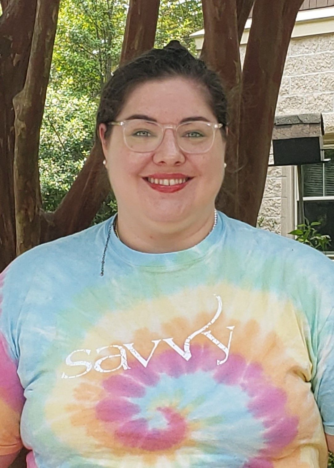 A woman with glasses and a red lip smiles at the camera while wearing a tie-dye shirt that says “savvy.”
