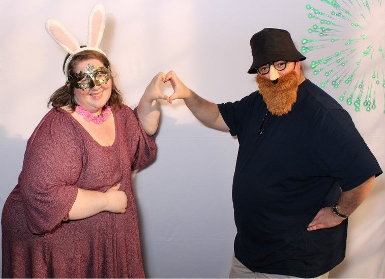 A woman in bunny ears and a masquerade mask and a man in a beard and hat, making a heart shape with their hands.