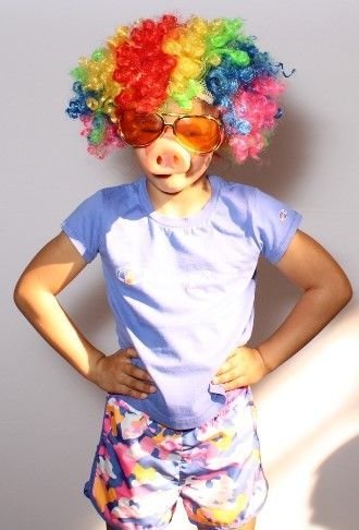 Child wearing a rainbow wig, orange-tinted glasses, and a pig nose, posing with hands on hips.