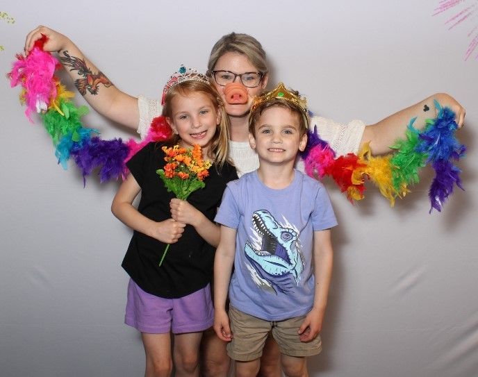 A woman with pig nose and wings, flanked by two children, all wearing crowns and smiling in front of a white backdrop.