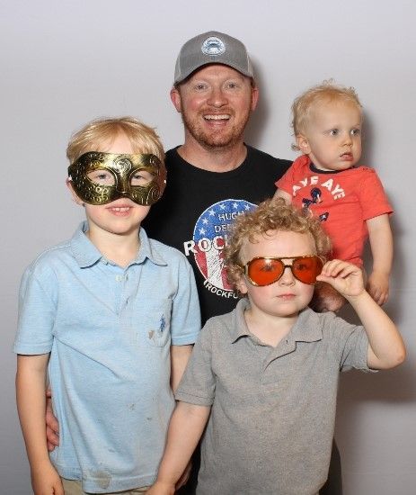 A man smiles, holding a baby, and posing with two young boys wearing sunglasses and a mask against a white backdrop.