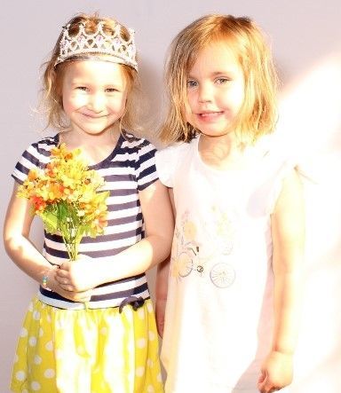 Two young girls smiling. One wears a tiara, striped shirt, and yellow skirt, holding flowers. The other wears a white shirt.