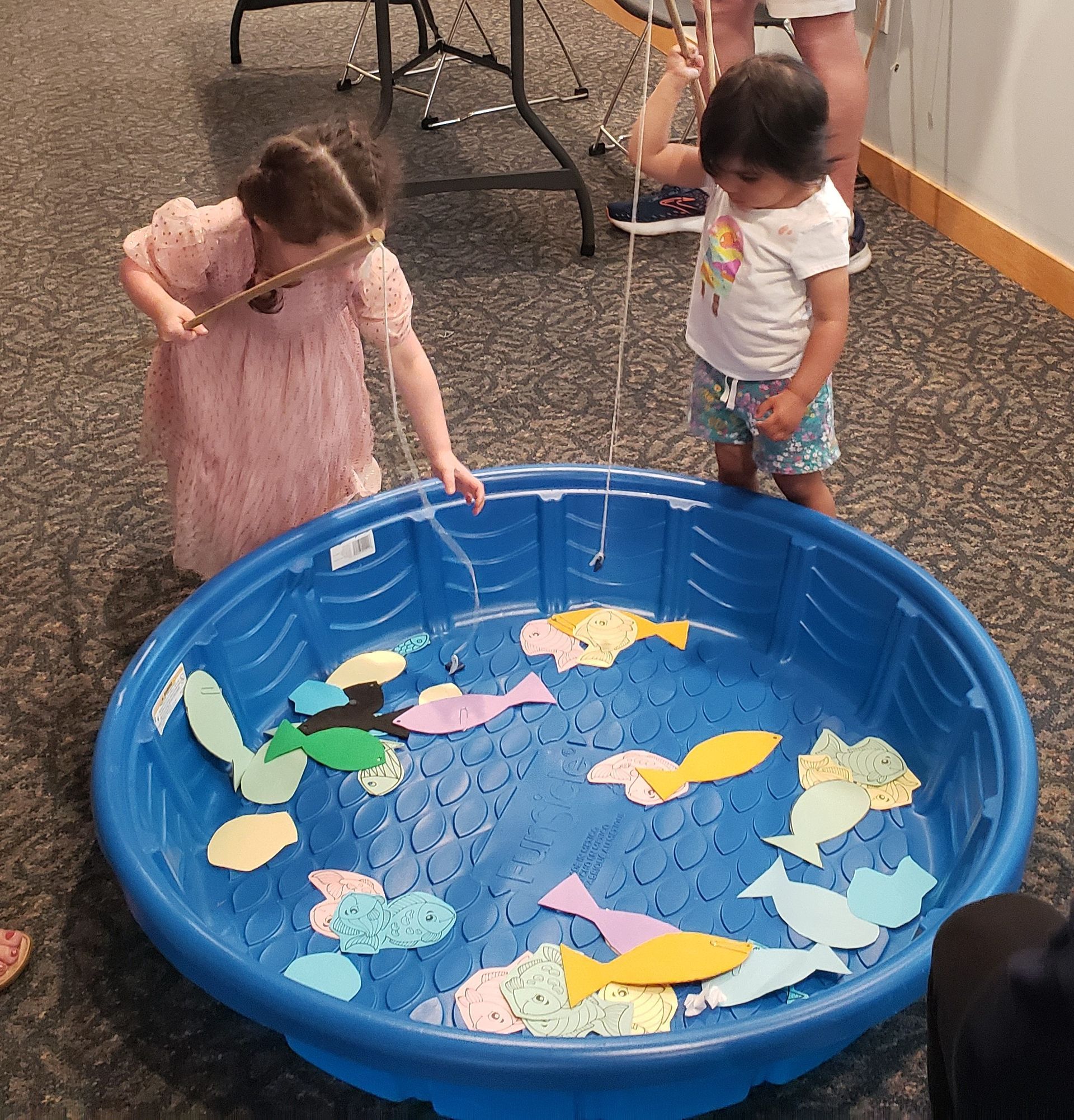 Two children fish for paper fish in a blue kiddie pool. One child is wearing pink, and the other a white shirt and blue shorts.