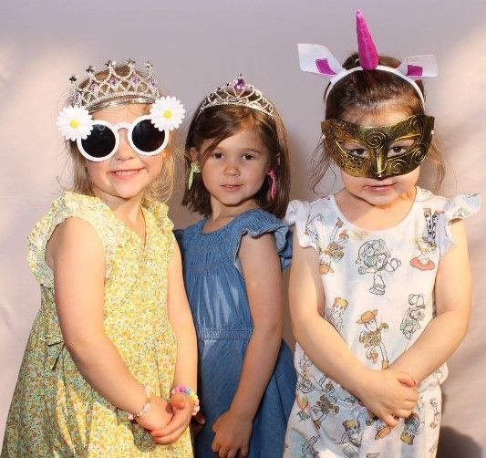 Three young girls in party attire. One wears daisy sunglasses and a crown, another a tiara, and the last a unicorn horn and mask.