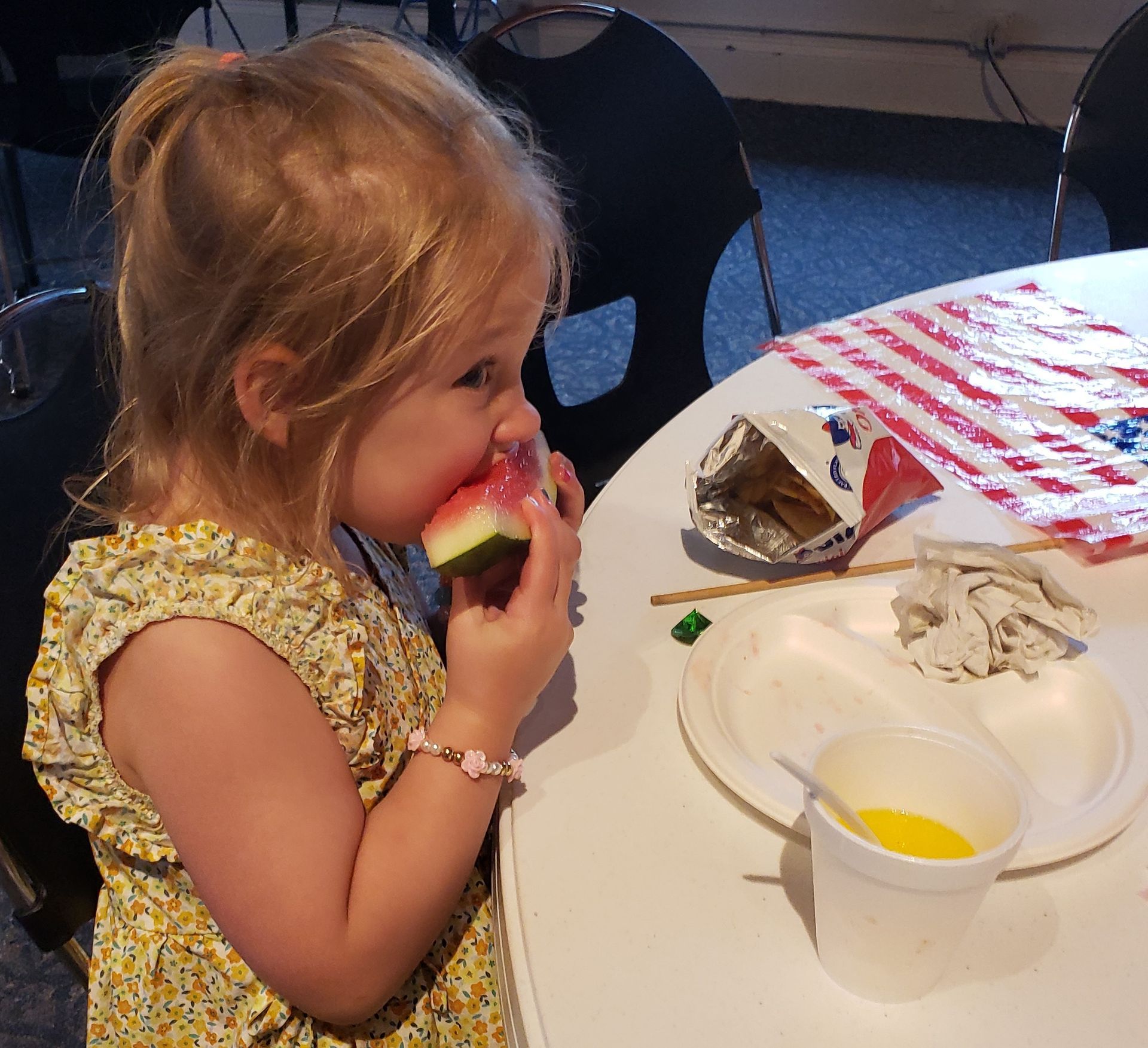 A young girl eats watermelon at a table. She wears a floral dress and has a bracelet. There's a flag and food on the table.