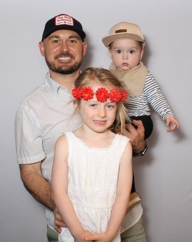 Man holding a baby and standing with a young girl. The man wears a baseball cap. The girl has a floral crown. Neutral background.