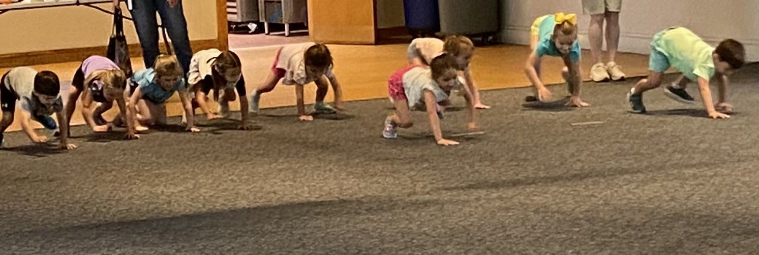 A group of children on a gray carpet are crawling in a line. One child is using a frisbee-like object to crawl on.