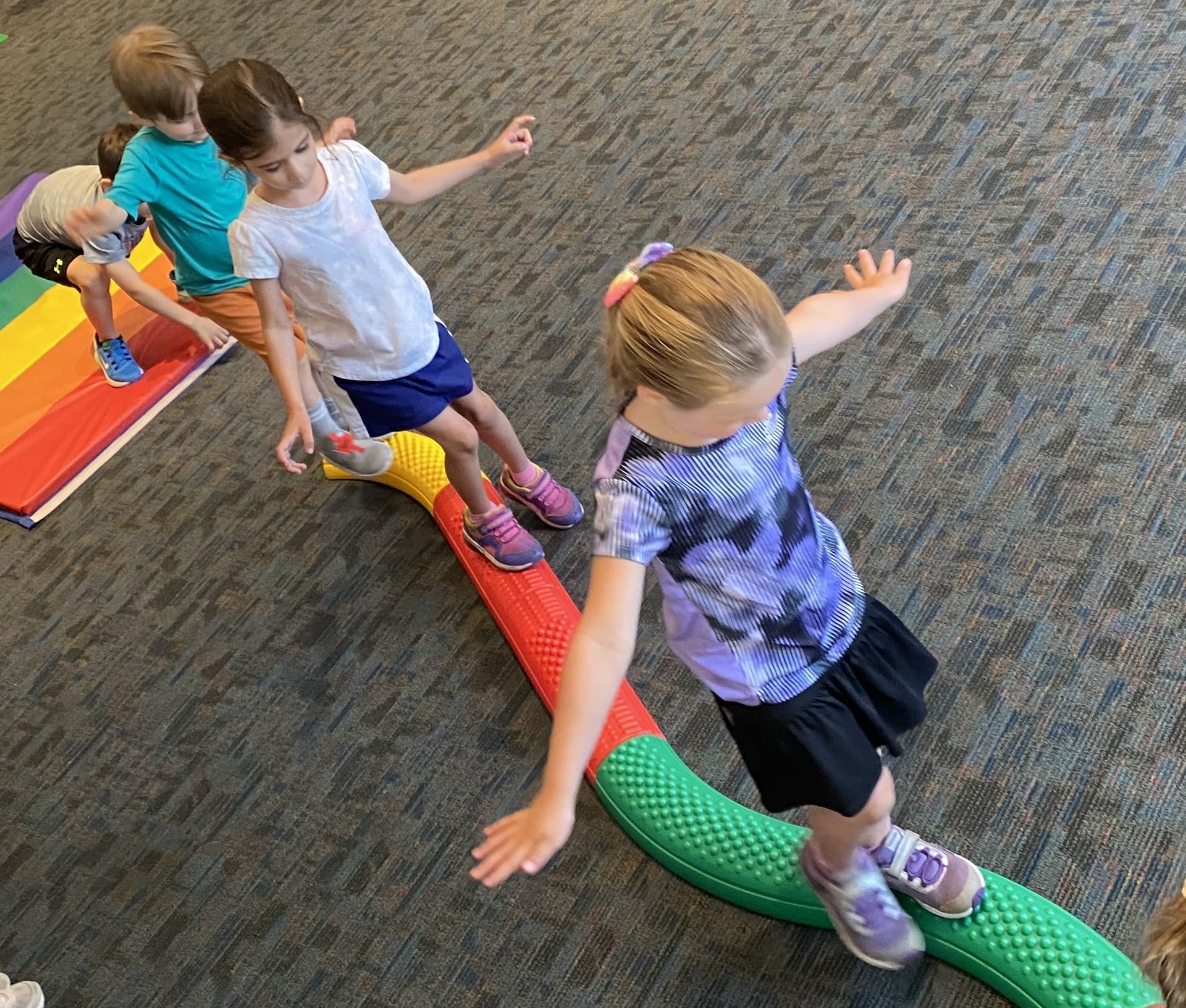 Children balancing on a colorful, curved balance beam indoors. They have arms outstretched for balance; one is wearing a tie-dye shirt.