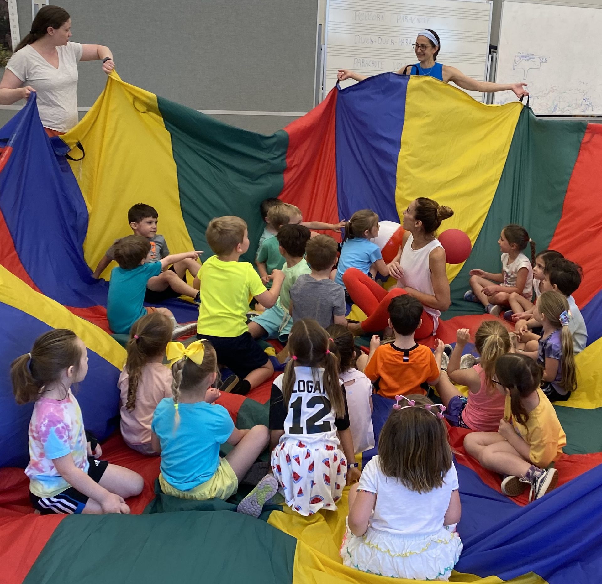 Children playing with a colorful parachute, led by three adults in a gymnasium.