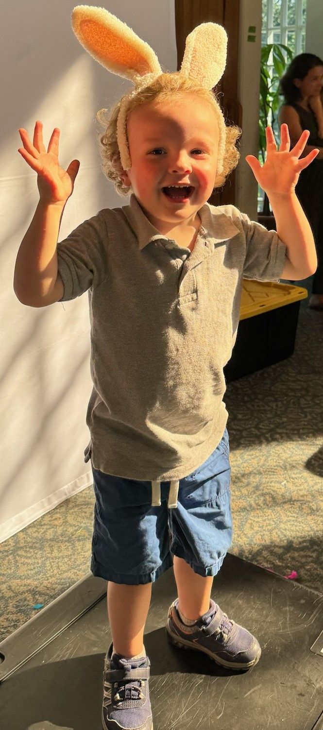 A young child wearing bunny ears, arms raised, smiling. They're in a gray polo shirt, blue shorts, and sneakers.