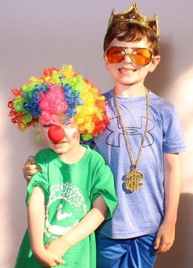 Two young children in costumes: One is a clown with a rainbow wig and red nose. The other wears a gold crown, sunglasses, and a gold chain.