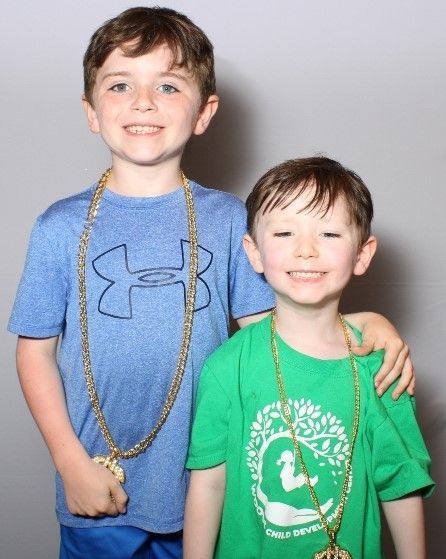 Two young boys wearing gold chain necklaces, smiling, and posing; the taller one has his arm around the other.