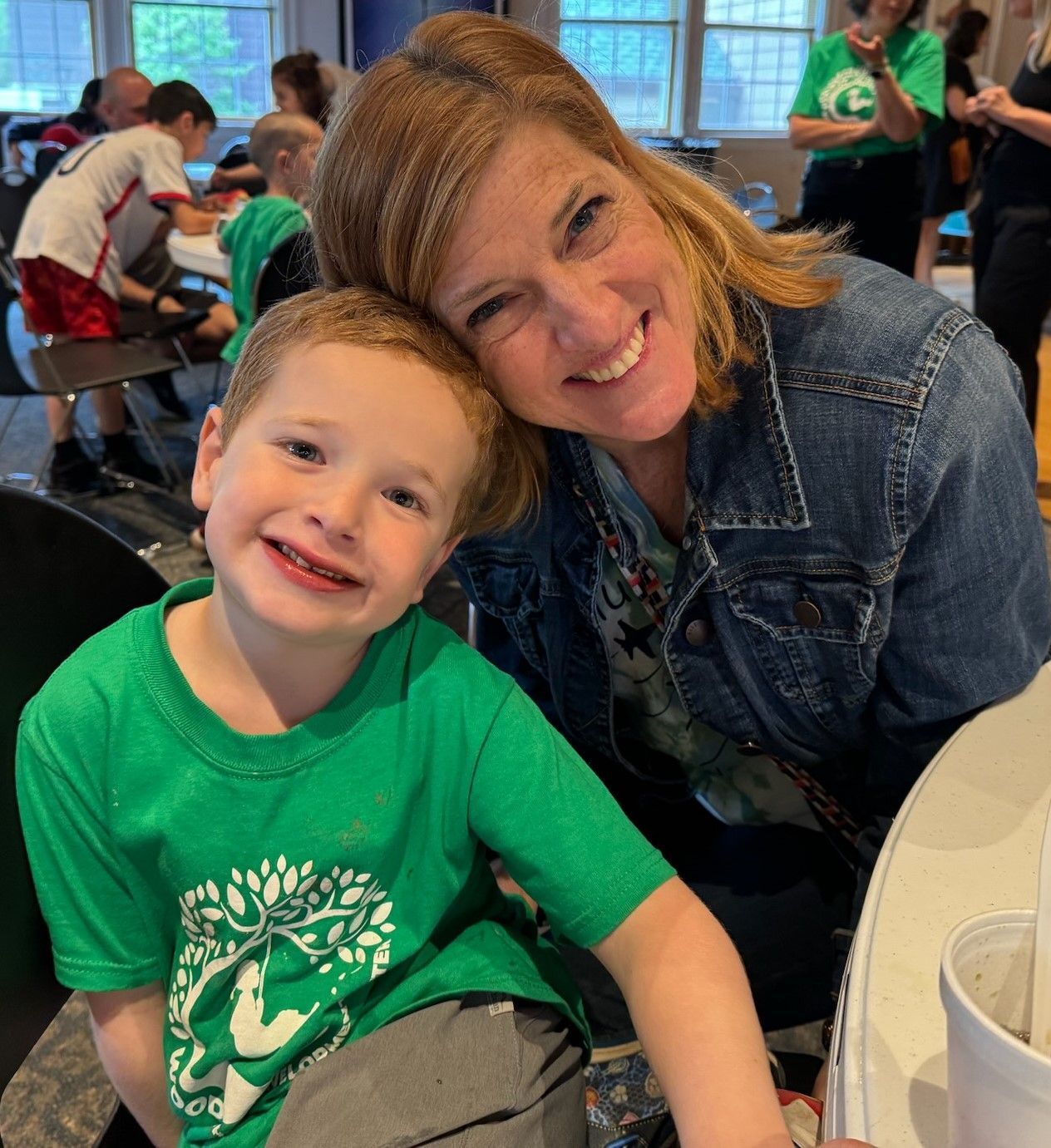 Woman in denim jacket smiles next to a smiling young boy in a green shirt. They sit at a table inside.