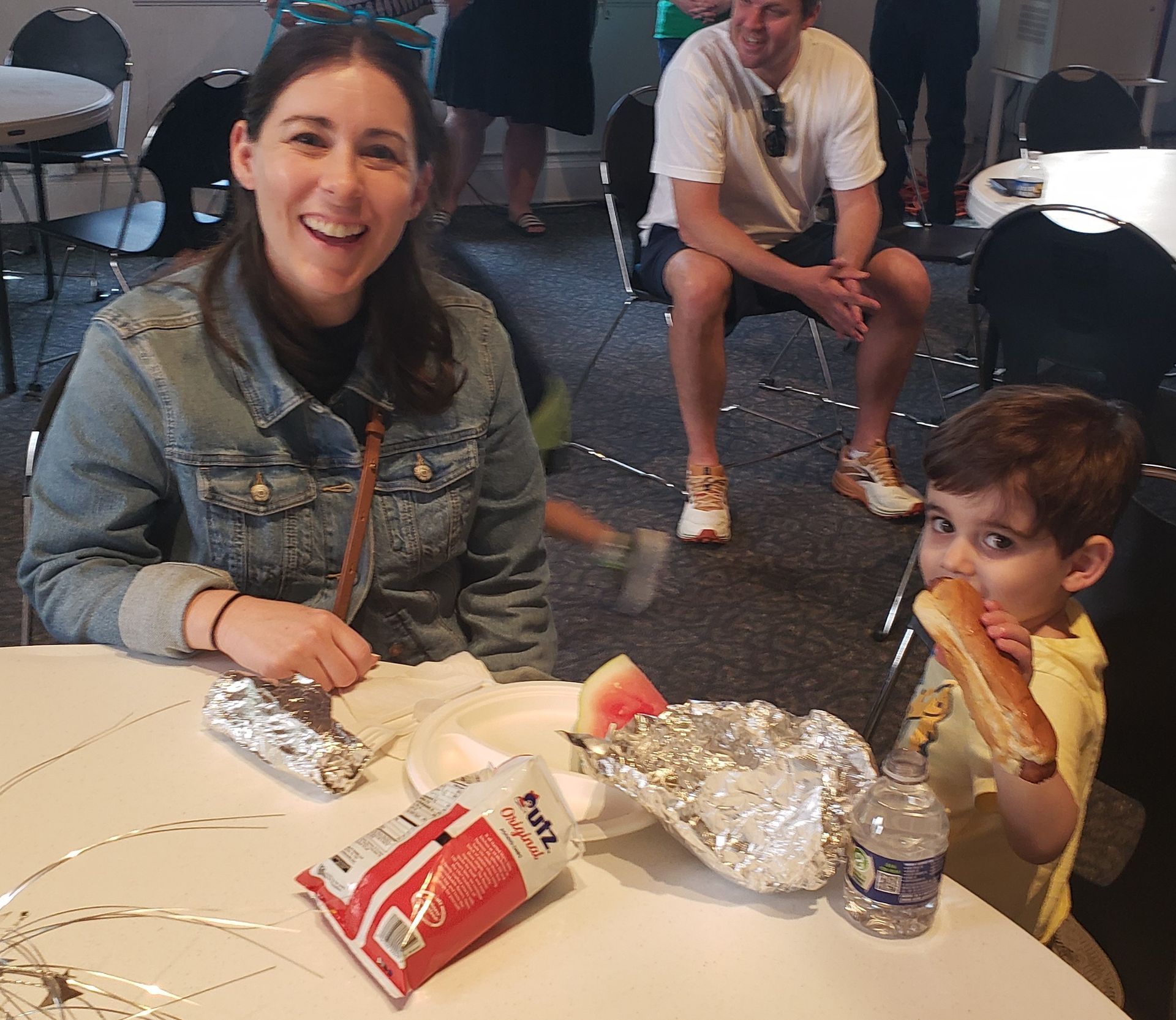 Woman and child at a table eating food, including a hot dog. The woman smiles broadly, while the child eats the hot dog.
