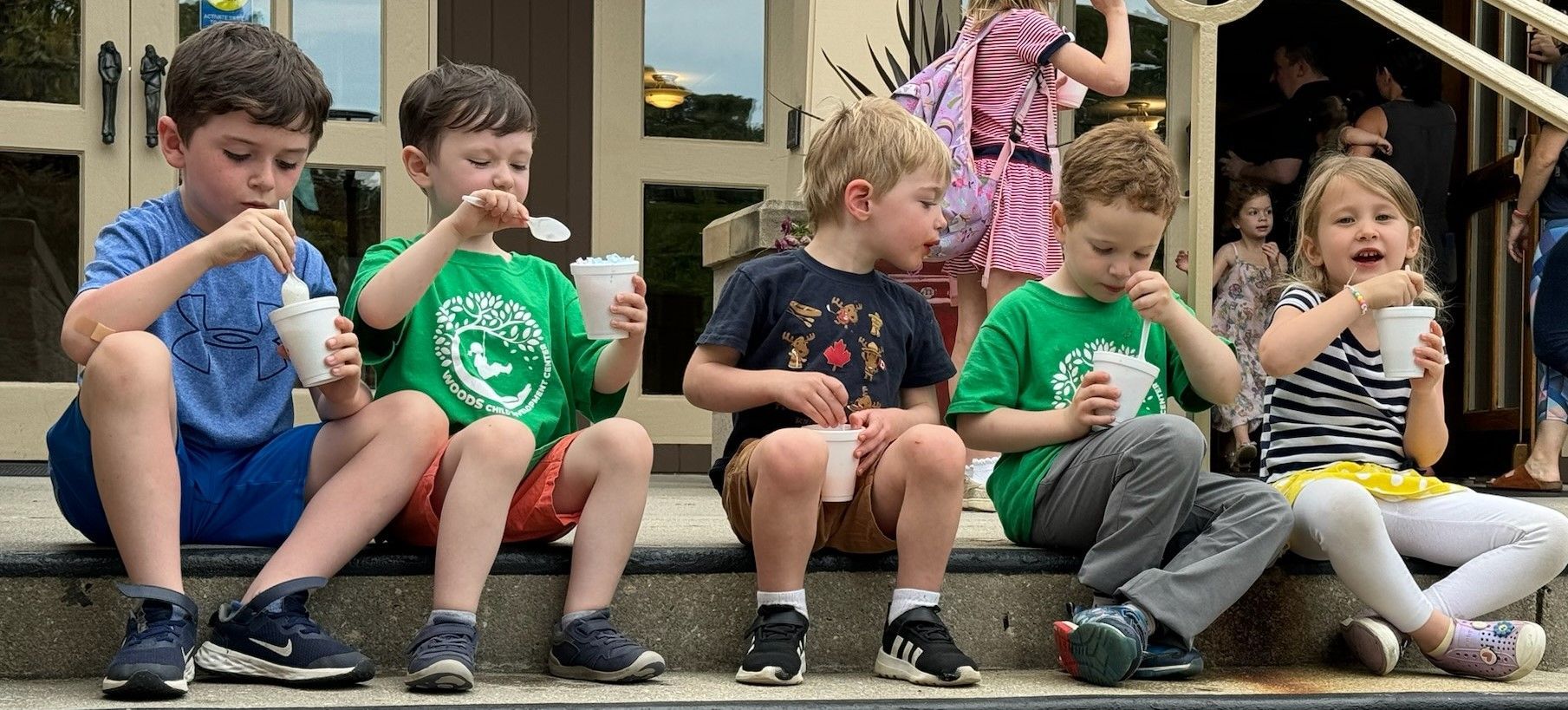 Six young children eating ice cream while sitting on steps outside. They wear various colored shirts and shorts, with excited expressions.