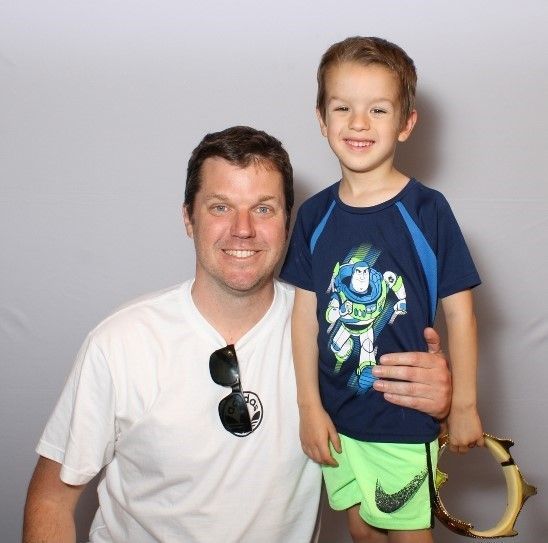 A man and young boy pose smiling. The boy wears a Buzz Lightyear shirt, green shorts, and holds a tambourine.