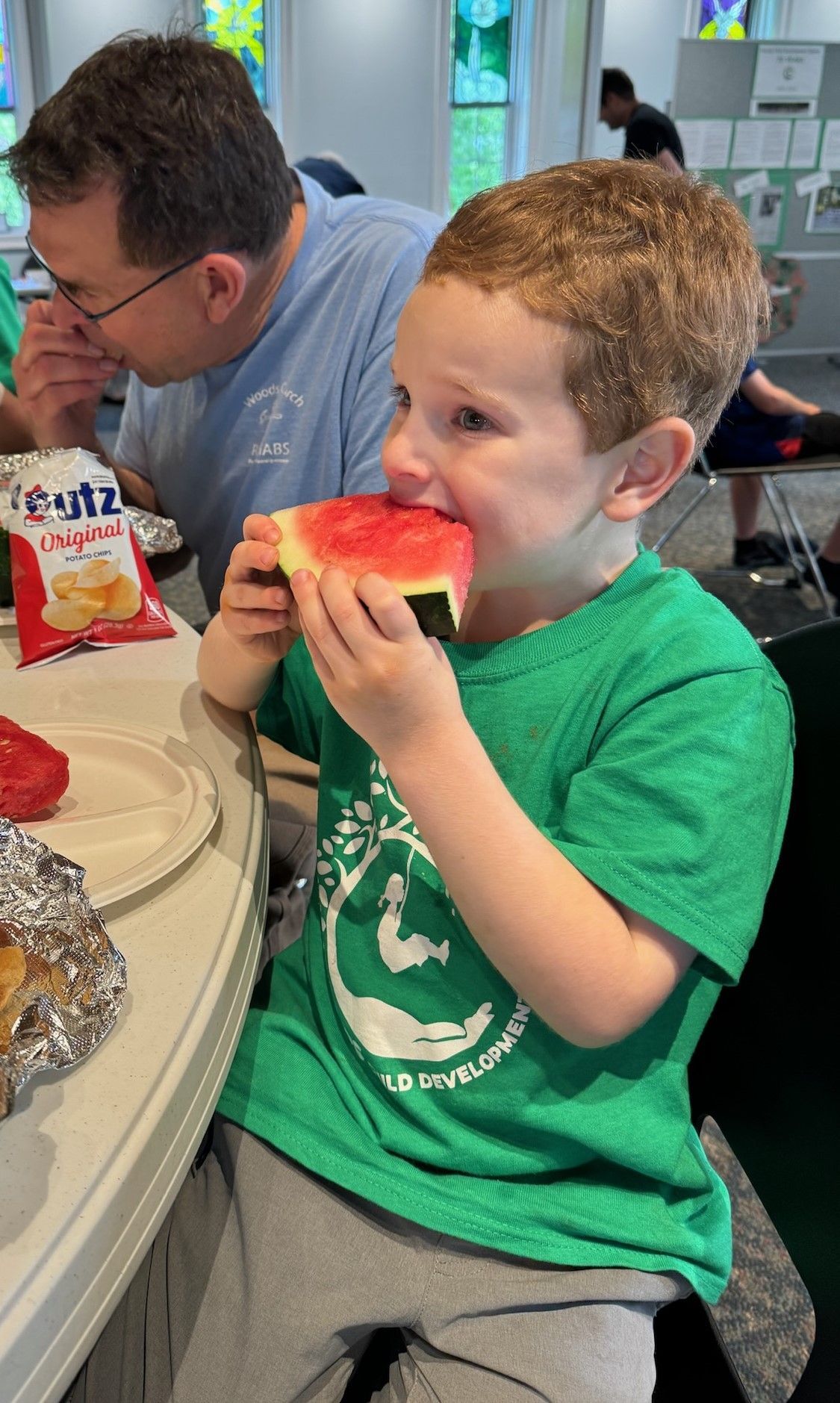 A young boy in a green shirt eats a piece of watermelon. He sits at a table indoors, with another person and snacks nearby.