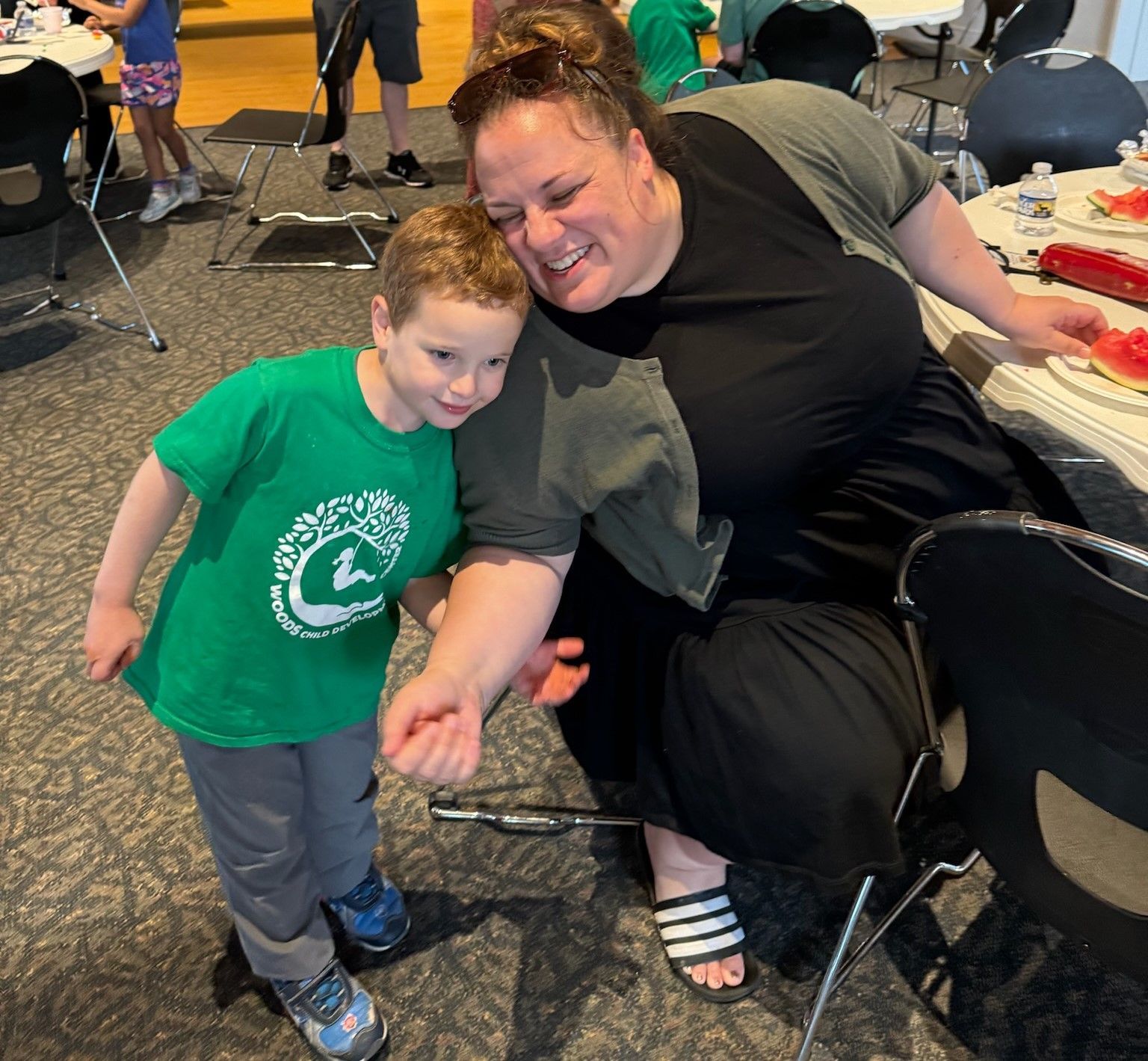 A woman with dark hair smiles at a young boy in a green t-shirt. They're looking at the boy's arm. Indoors with other people.