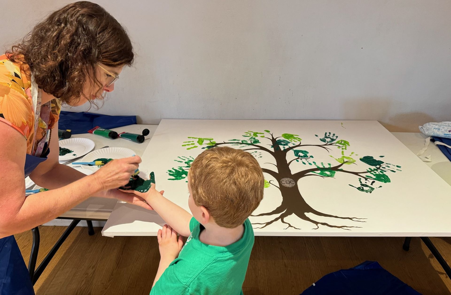 Woman helping a young child with a handprint art project on a large tree outline. They are indoors at a table.