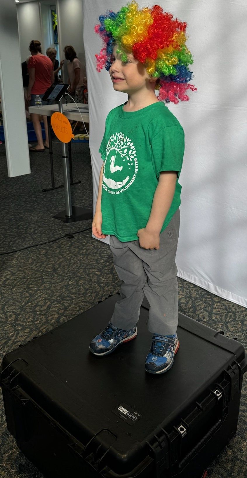 A young boy in a rainbow clown wig and green shirt stands on a black box. He is in a room with other people.