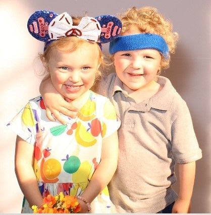 Two young children smiling: a girl wearing a fruit-print dress and a Mickey Mouse ear headband, and a boy wearing a gray shirt and a blue headband, with his arm around the girl.