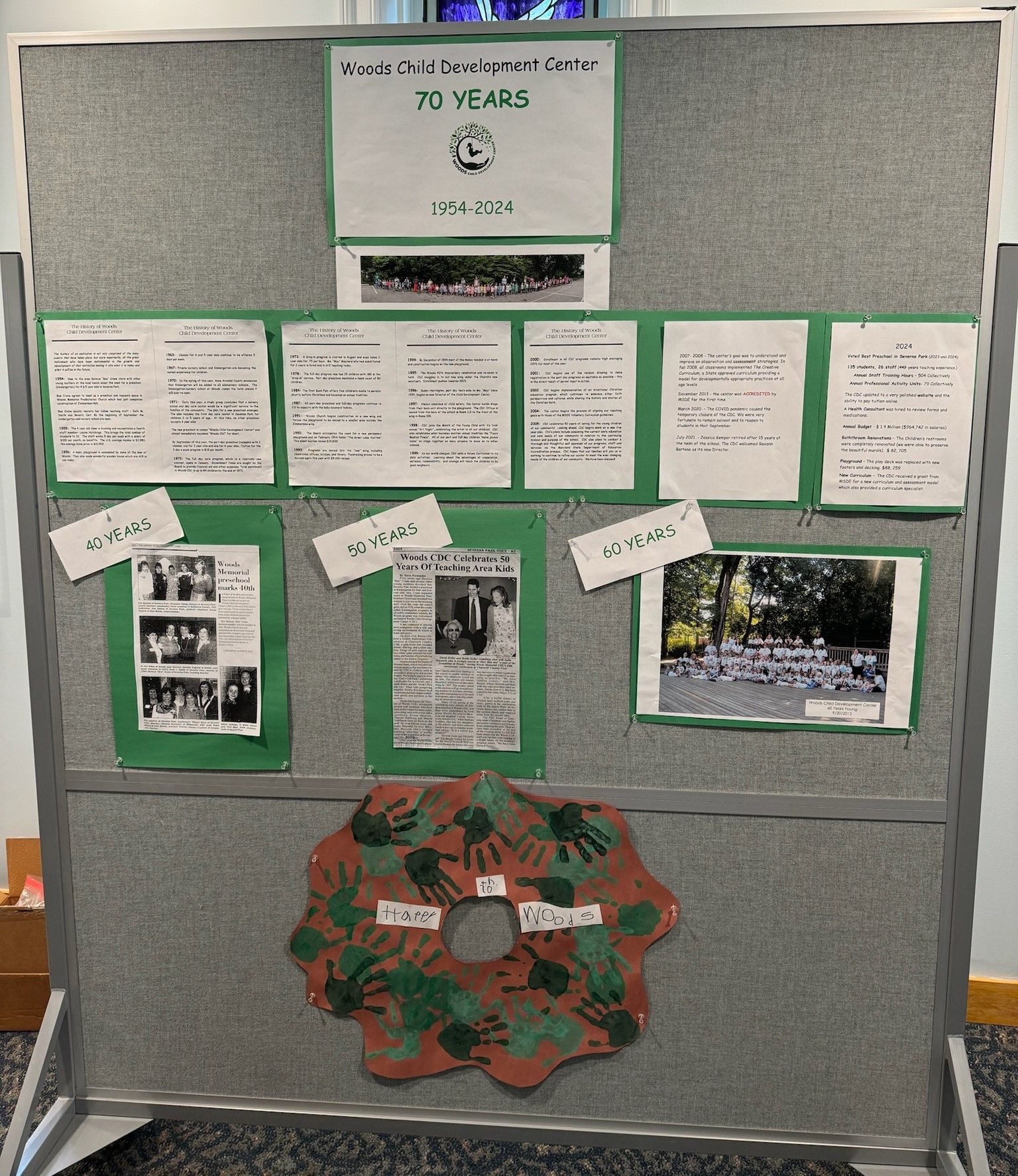 A display board celebrating a 70th anniversary, featuring photos, text, and a handprint wreath. The board has a gray background with green accents.