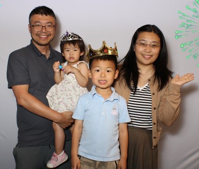 A family of four smiling. The father holds a young child wearing a tiara, the older child wears a crown, and the mother gestures.