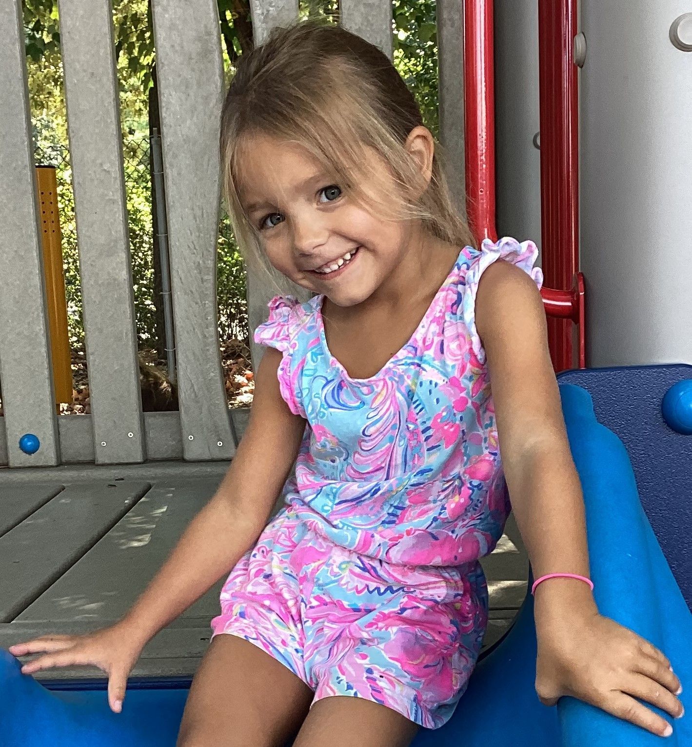 Young girl in a pink and blue romper smiles at the camera, sitting on blue playground equipment.