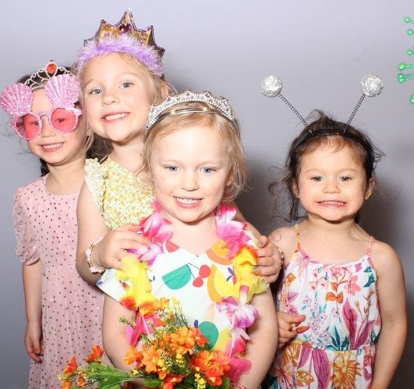 Four young girls posing for a photo booth. They wear party accessories and smile, with floral lei and crown visible.