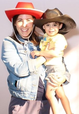 Woman in a denim jacket, wearing a red cowboy hat, holds a young child in a brown cowboy hat; both smile.