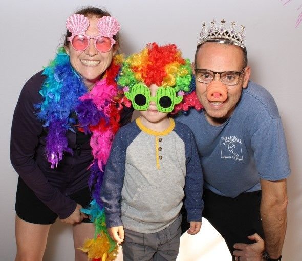 Family posing in a photo booth with funny props: woman with seashell glasses and boa, child with clown wig and green glasses, man with pig nose and crown.