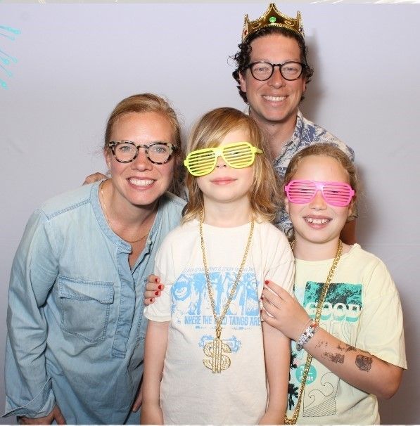 A family of four in a photo booth. Two children wear novelty glasses and gold chain necklaces, the dad wears a crown, and everyone smiles.